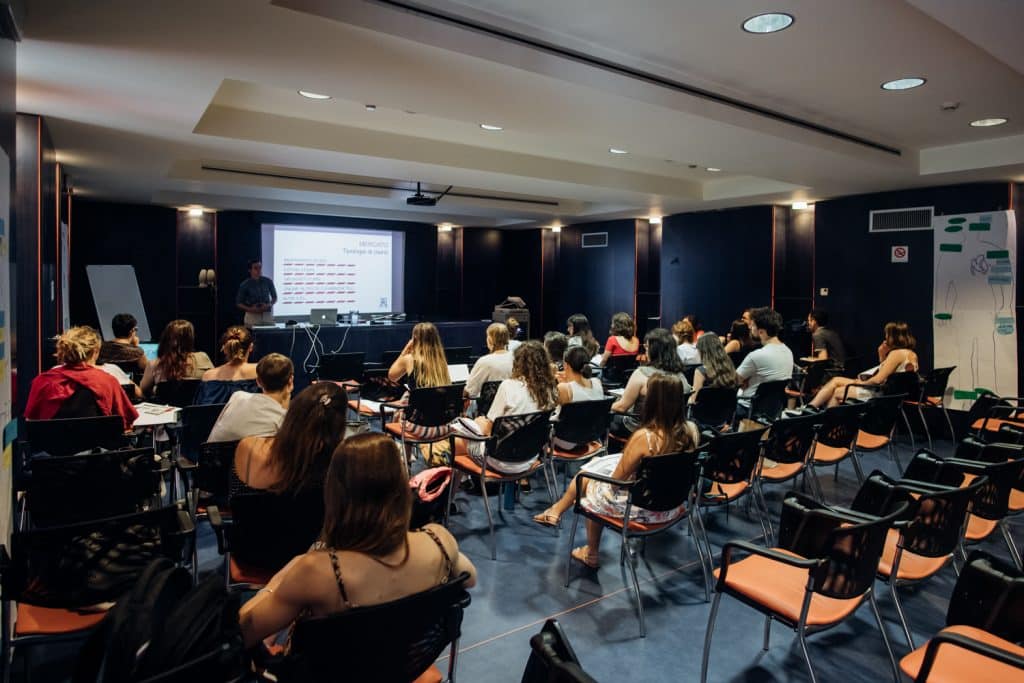 gruppo di studentesse e studenti seduti su sedie ascoltano il professore mentre spiega